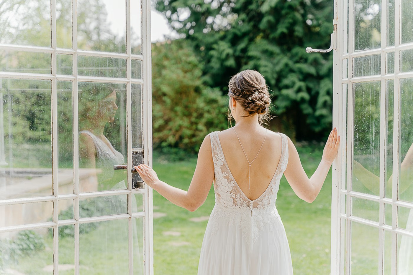 Brautkleid Schlosspark Destedt – Braut steht von hinten in einer ge&ouml;ffneten Glas-Fl&uuml;geltür des Palmengartens und blickt in den gr&uuml;nen Park, stille Vorfreude, Hochzeitsfotografie Schlosspark Destedt Kathrin Skorpel