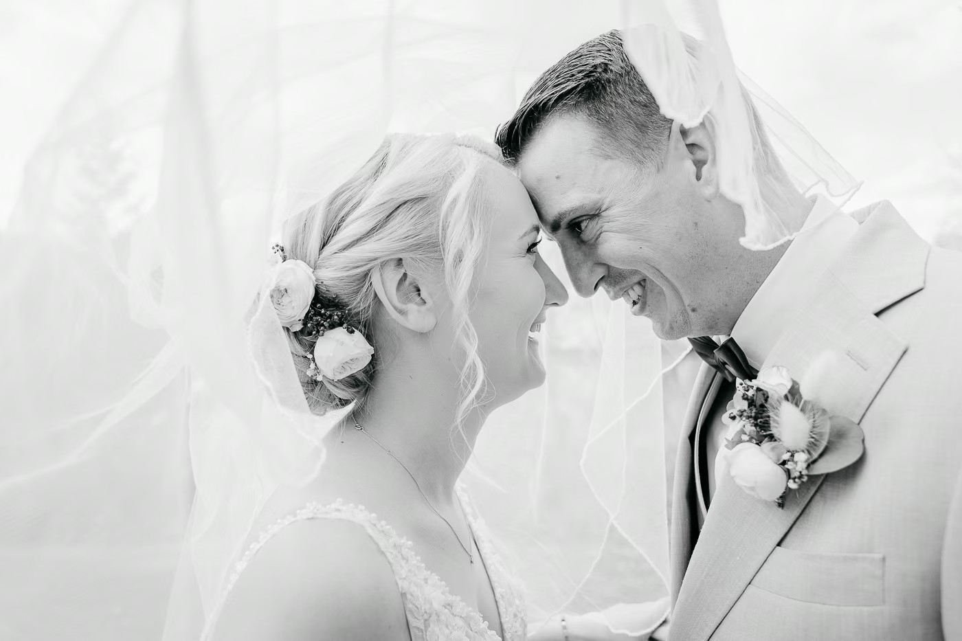 Paarshooting Meine – Schwarz-Weiß-Aufnahme, Brautpaar lacht Stirn an Stirn unter wehendem Schleier, Blumenschmuck im Haar der Braut, verspielte Ausgelassenheit, Hochzeitsfotografie Meine Kathrin Skorpel