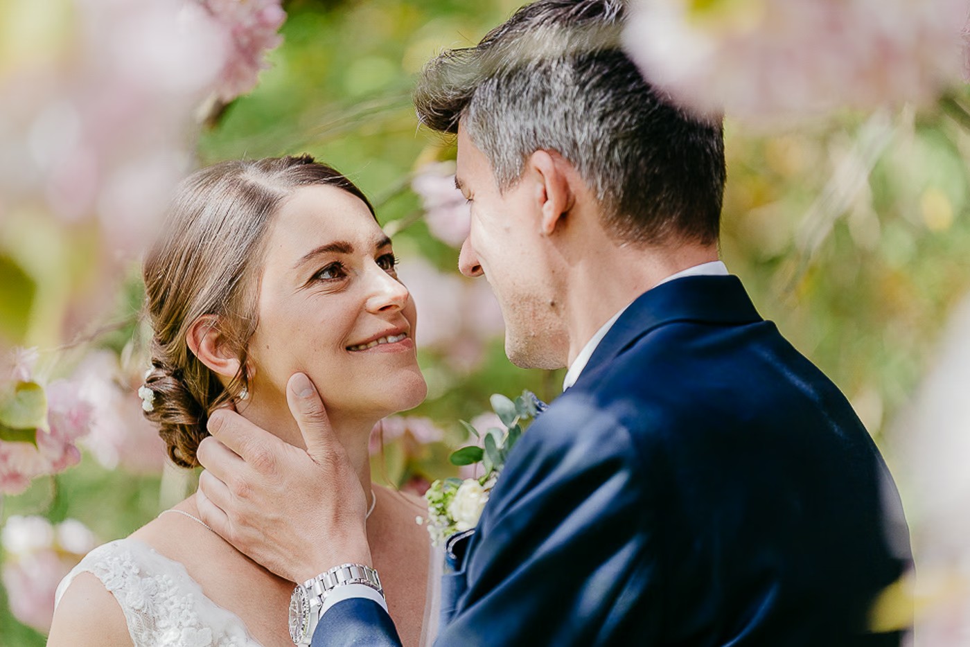 Paarshooting Schlosspark Cremlingen – Bräutigam hält zärtlich das Kinn der Braut unter blühenden rosa Zweigen, inniger Blickkontakt, romantische Frühlingsstimmung, Hochzeitsfotografie Schlosspark Cremlingen Kathrin Skorpel