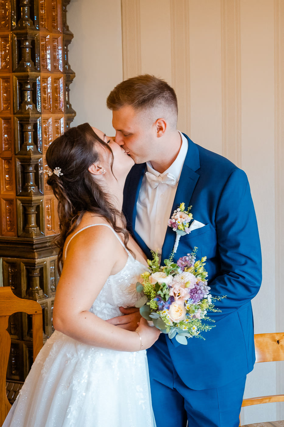 Standesamt Hochzeit Helmstedt – Brautpaar küsst sich vor historischem Kachelofen, Bräutigam im blauen Anzug hält Braut mit buntem Strauß im Arm, romantische Hochzeitsreportage Helmstedt Kathrin Skorpel