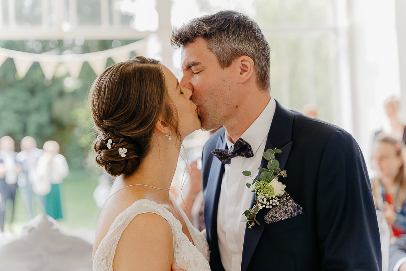 Standesamt Hochzeit Salzgitter – Brautpaar küsst sich beim ersten Kuss als Eheleute unter einer Wimpelkette im lichtdurchfluteten Pavillon, Gäste applaudieren im Hintergrund, Hochzeitsreportage Salzgitter Kathrin Skorpel